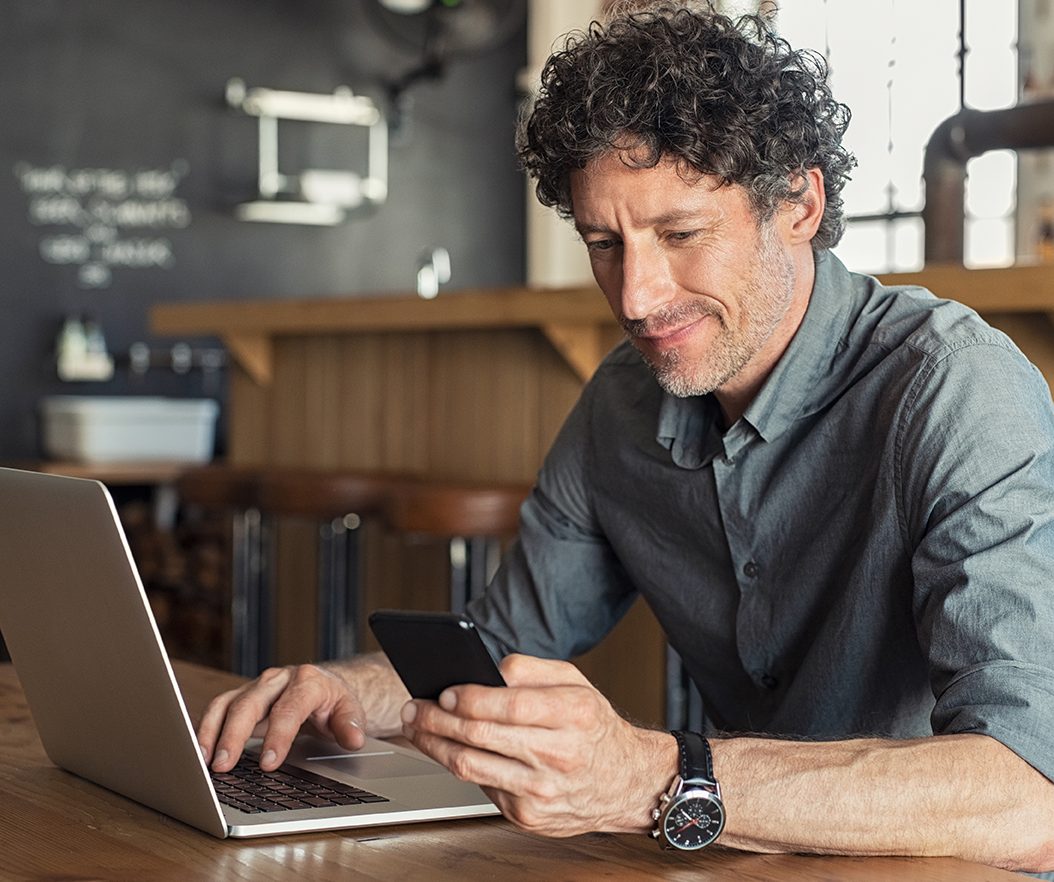 Older man sits at table in cafe on laptop and looking at phone